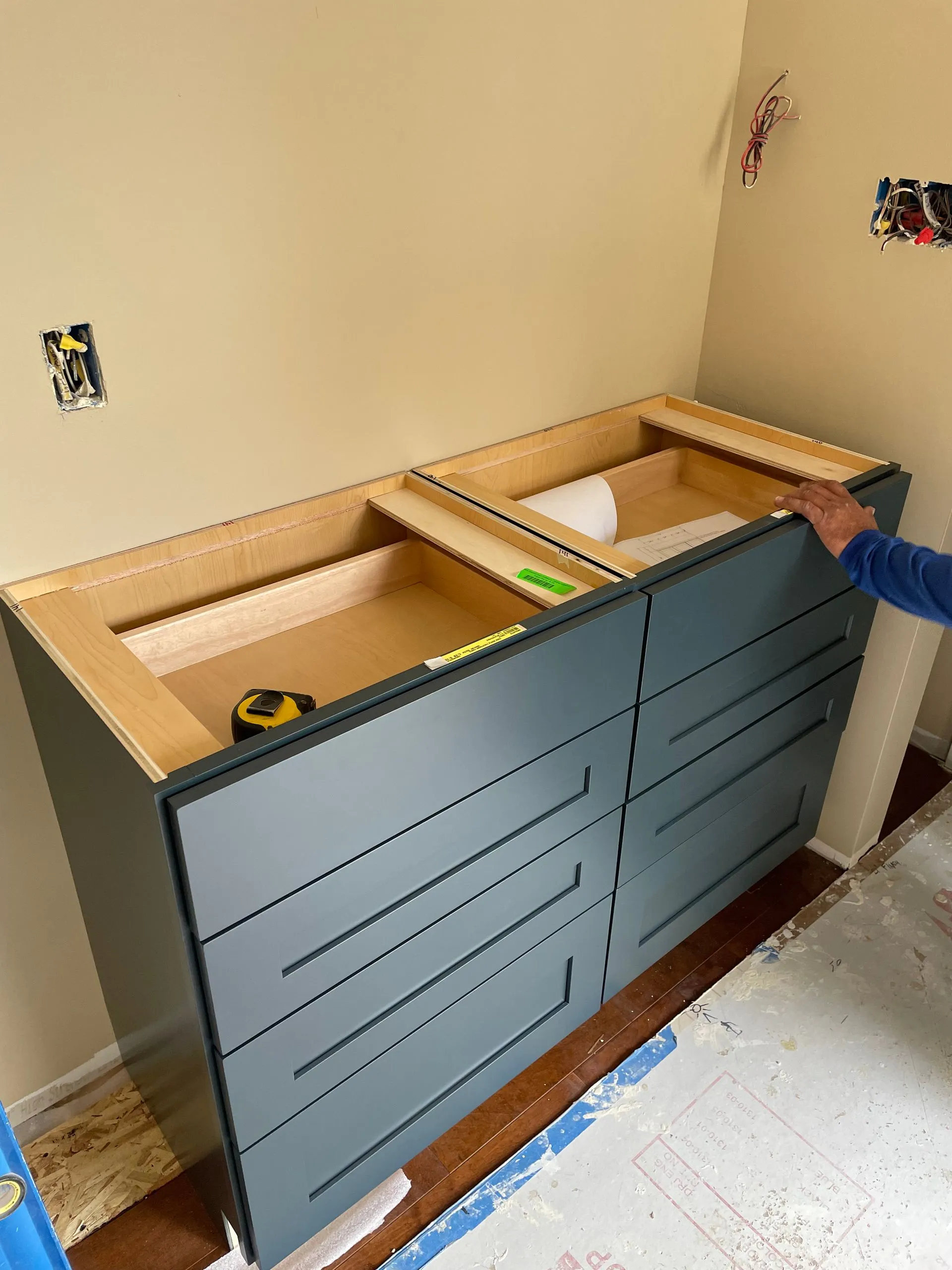 Blue vanity cabinet with open countertop cutouts under construction in a beige room, measuring tape inside.