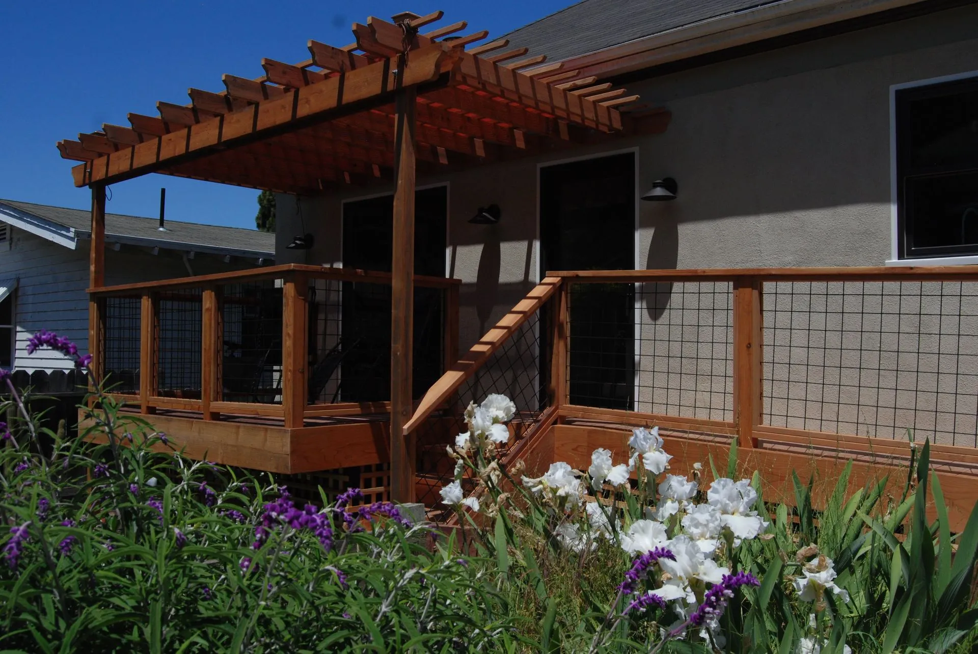 House deck with wooden pergola and railing, framed by white and purple flowers in a sunny yard.