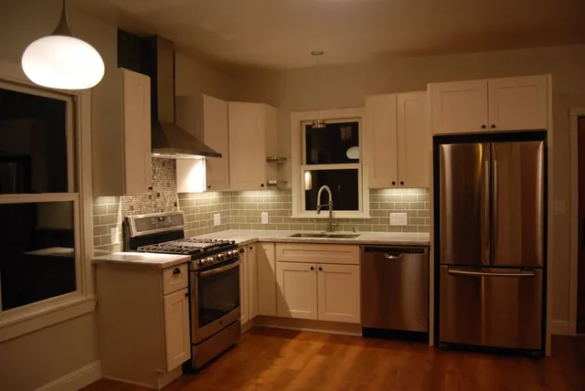 Modern kitchen with white cabinets, stainless steel appliances, and warm wood floors at night