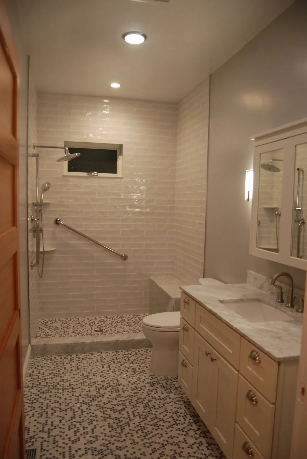 Narrow accessible bathroom with tiled walk-in shower, grab bar, toilet, and vanity under bright lights