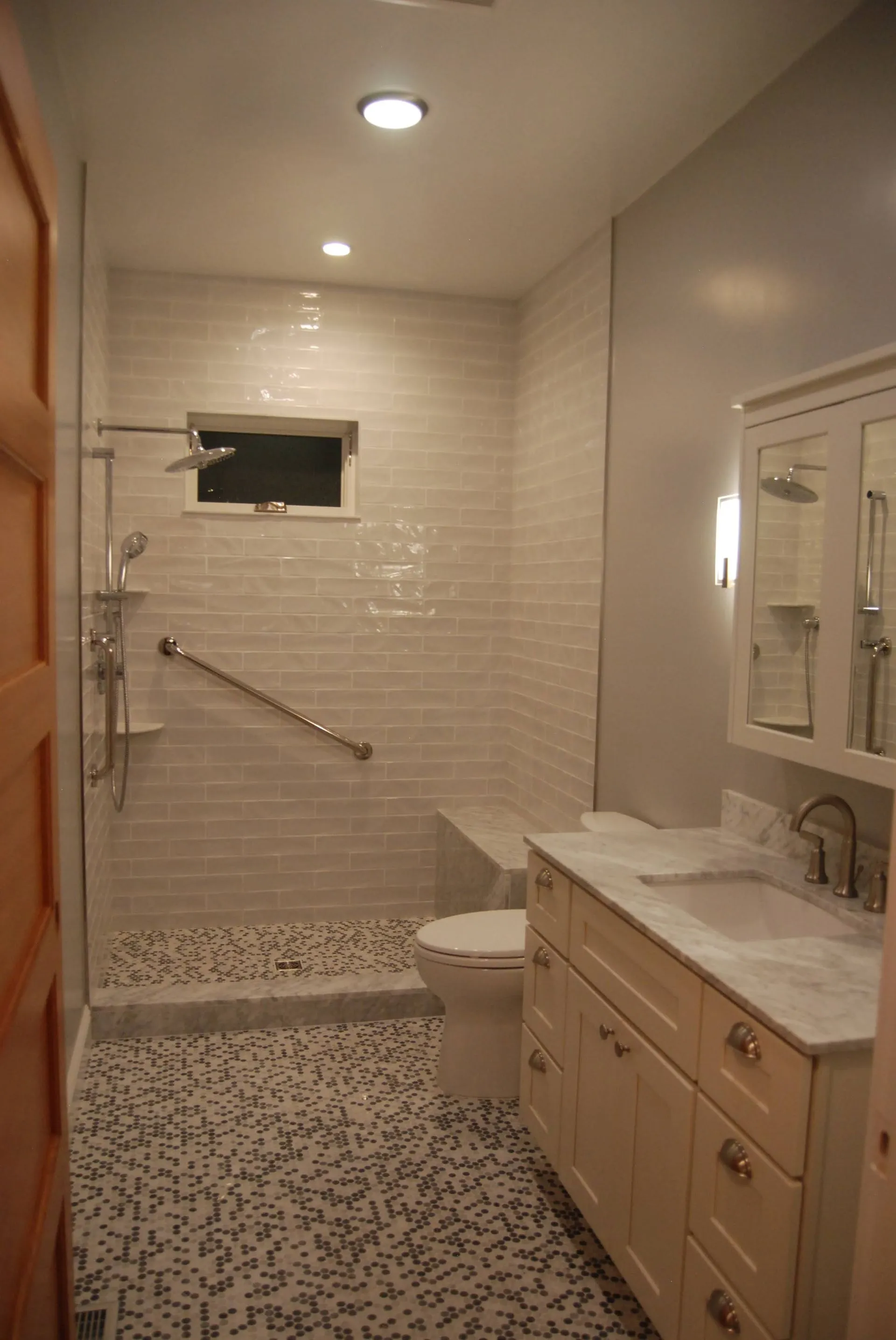 Narrow accessible bathroom with tiled walk-in shower, grab bar, toilet, and vanity under bright lights