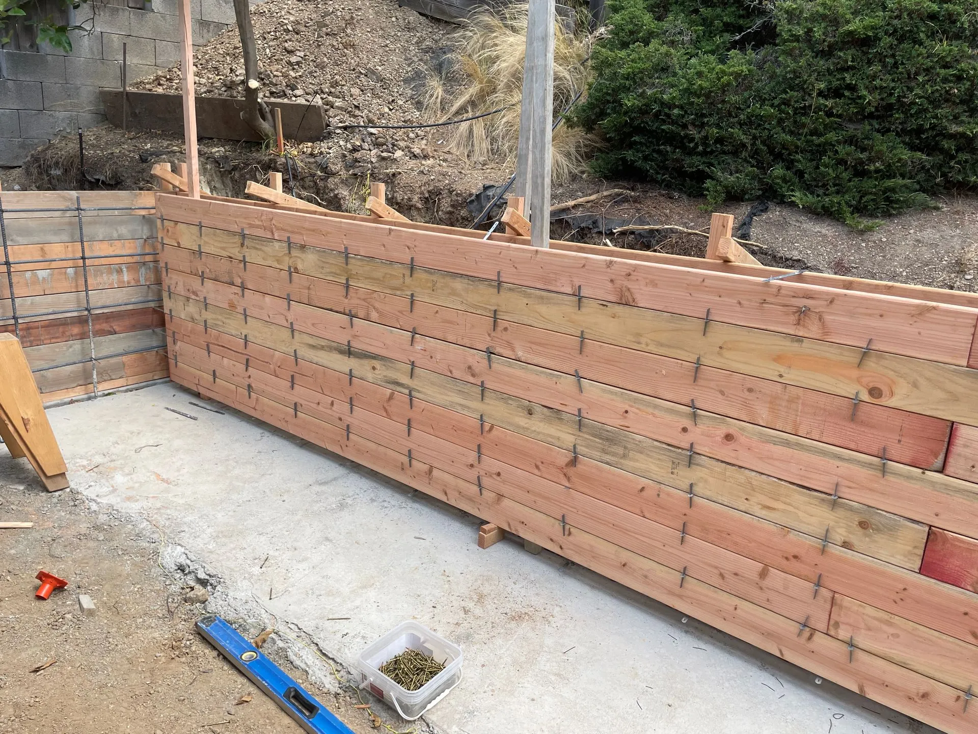 Wooden retaining wall under construction on a concrete foundation beside a dirt slope.