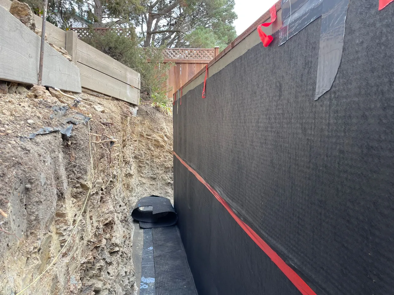 Narrow construction trench with rocky wall, black mesh fencing, and a drainage pipe along the bottom.