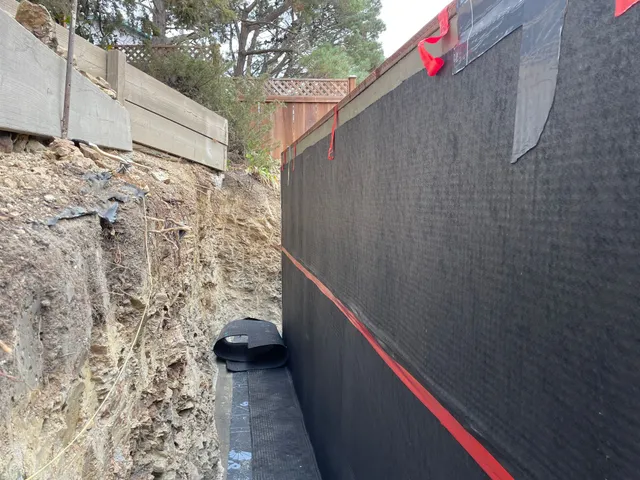 Narrow construction trench with rocky wall, black mesh fencing, and a drainage pipe along the bottom.