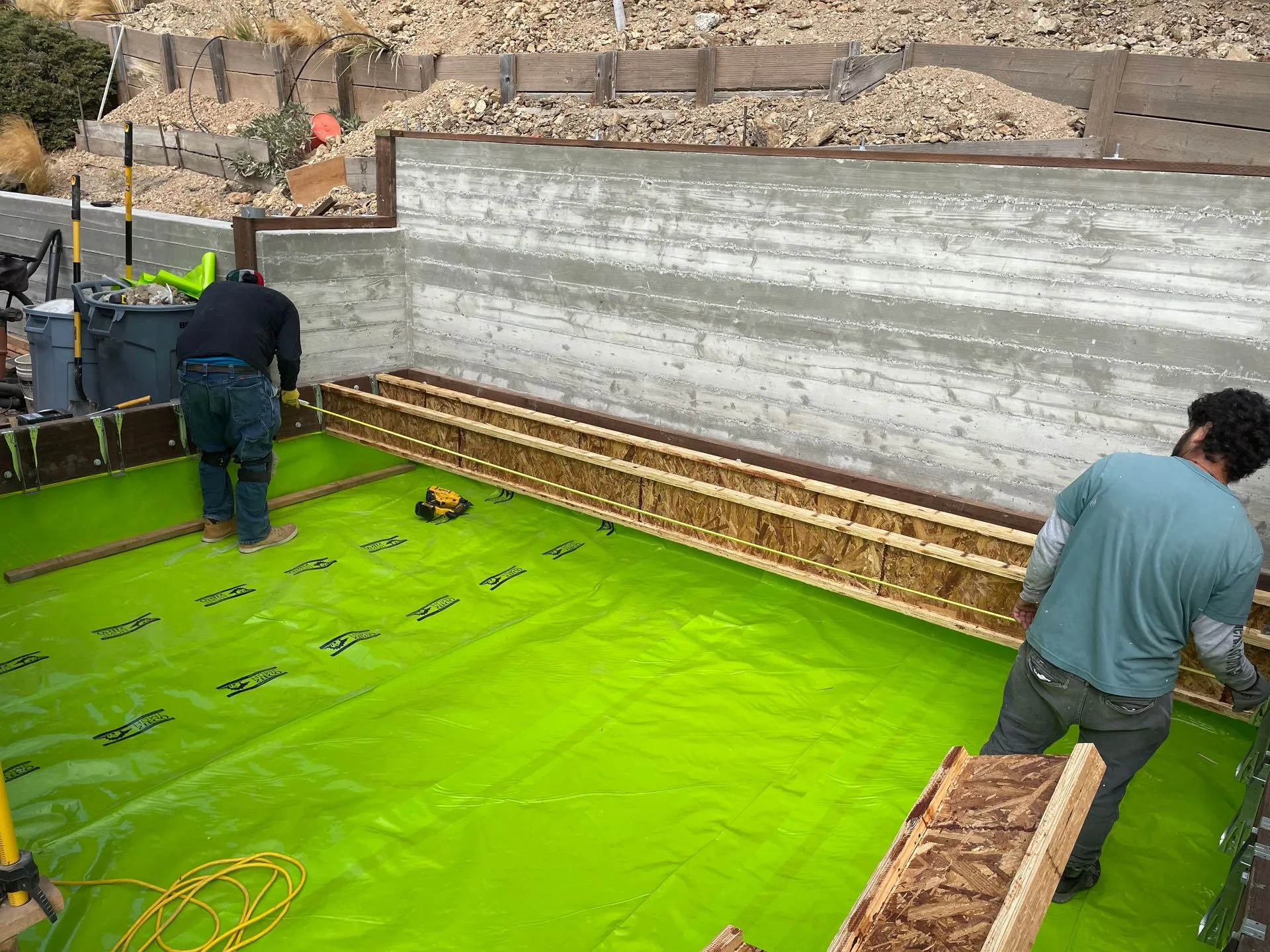 Workers installing bright green waterproofing on a construction foundation area.