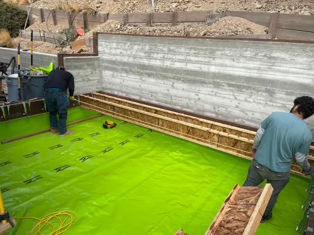 Workers installing bright green waterproofing on a construction foundation area.