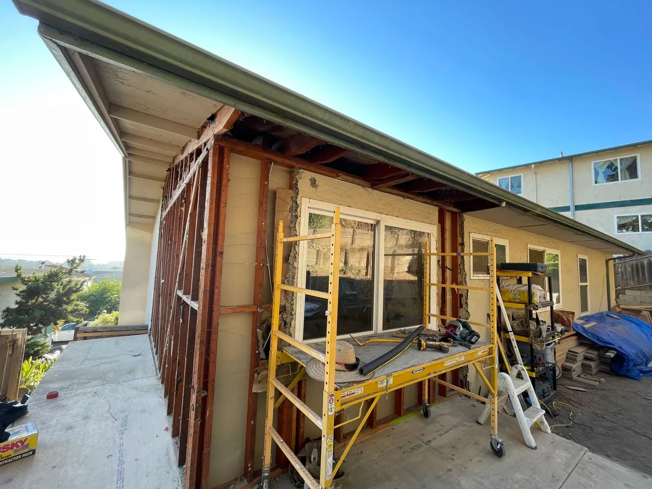 House exterior under renovation with scaffolding, ladders, and exposed framing beneath a sloped roof