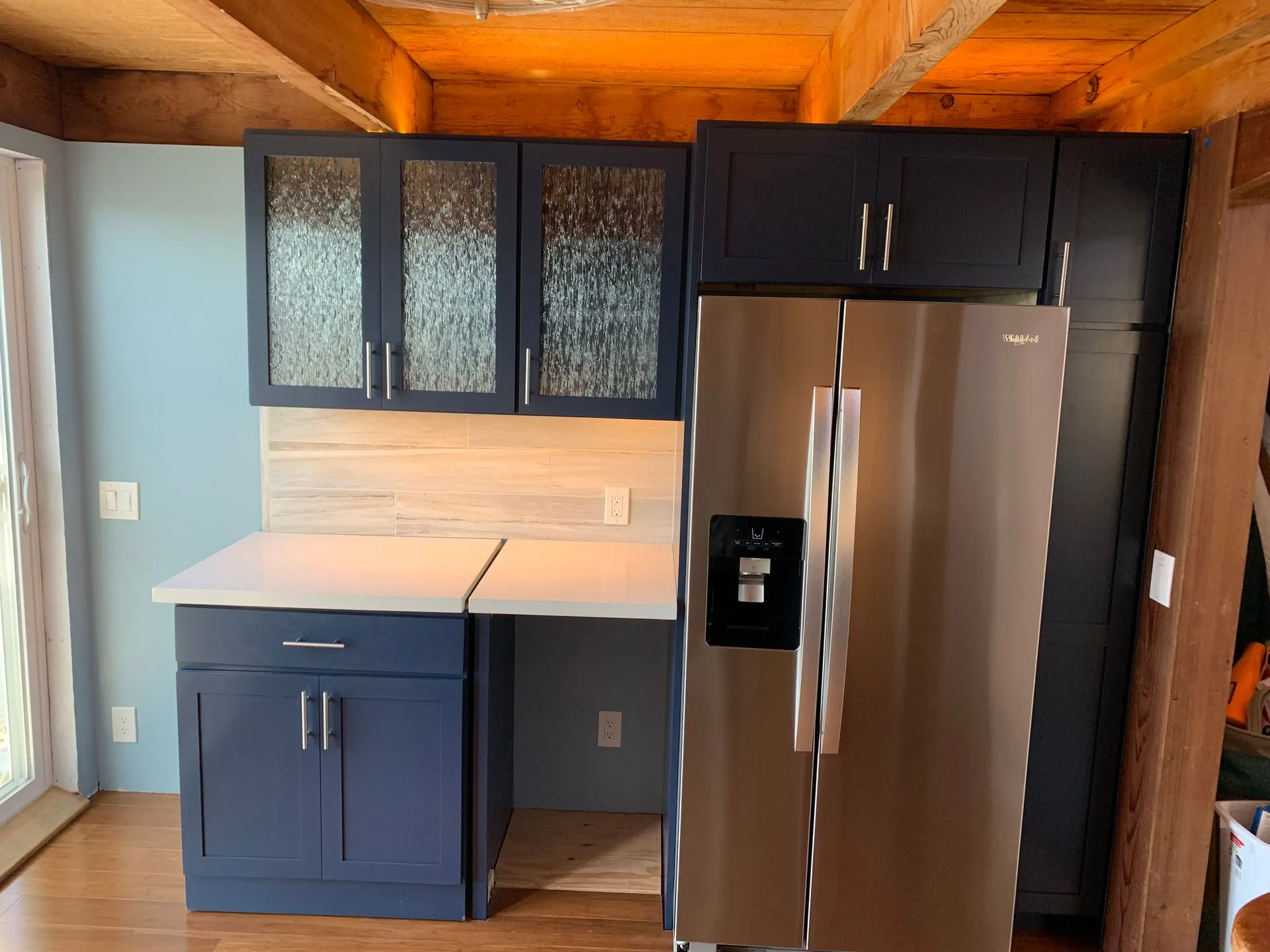 Modern kitchen corner with blue cabinets, white countertops, and stainless steel refrigerator under wood ceiling
