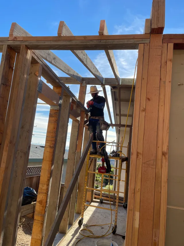 Worker on scaffolding inside a wooden building frame, wearing a harness and using a power tool.