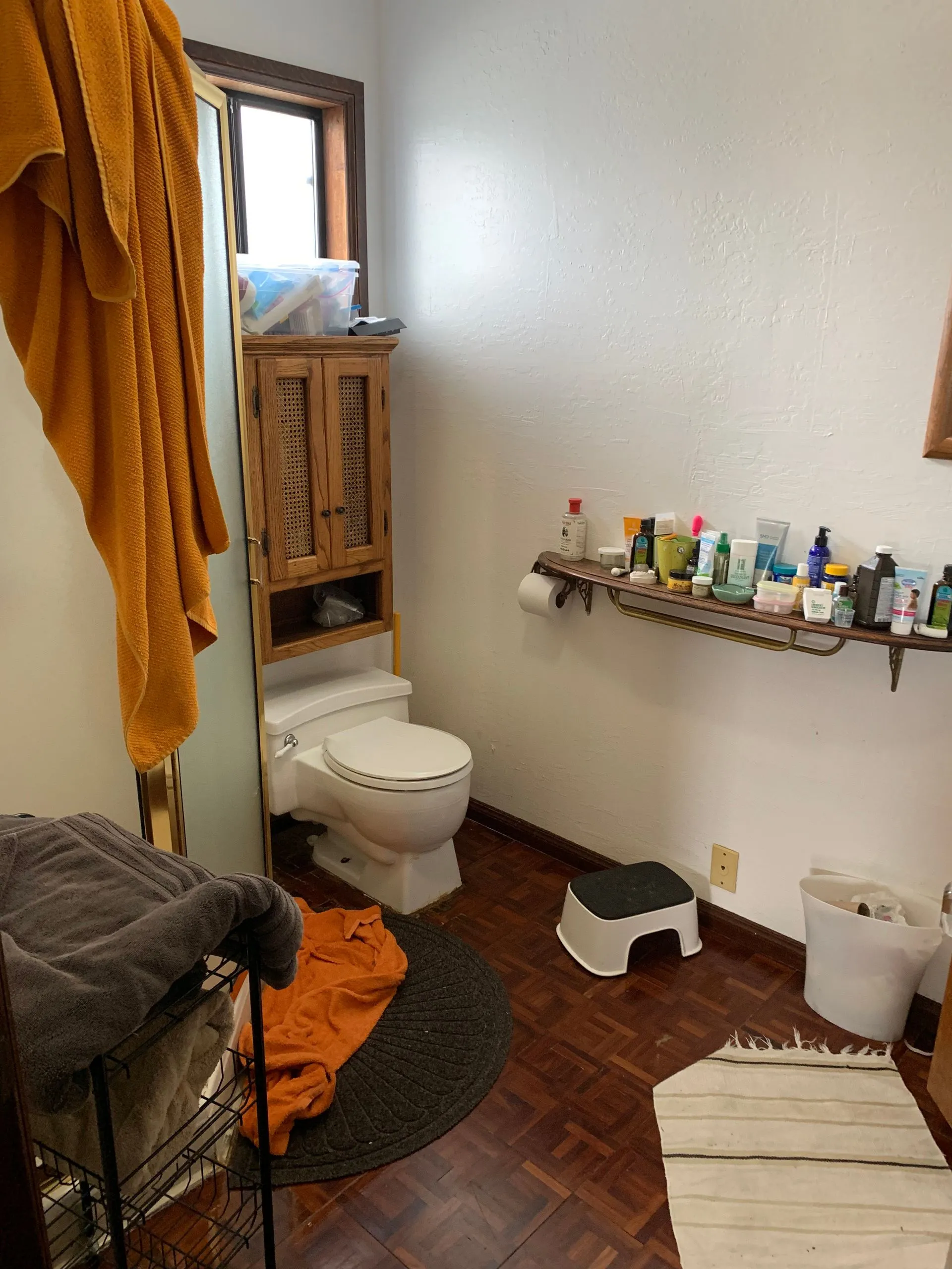 Small bathroom with toilet, towel rack, toiletries shelf, hamper, step stool, and striped rug.