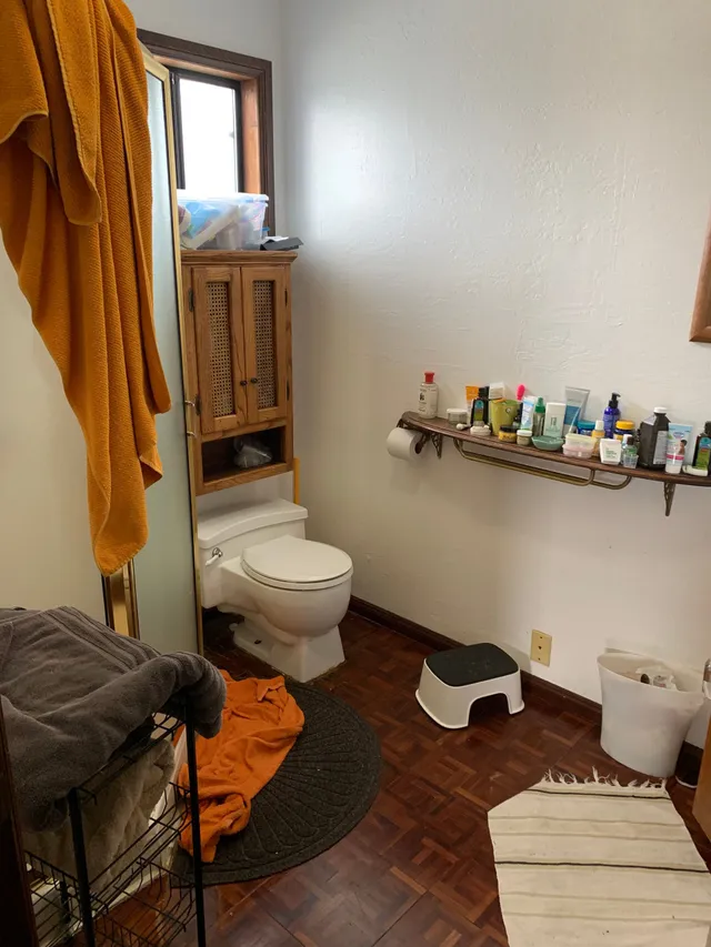 Small bathroom with toilet, towel rack, toiletries shelf, hamper, step stool, and striped rug.