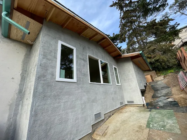 Gray stucco house exterior with white-framed windows, wooden eaves, and a narrow concrete side yard by stairs