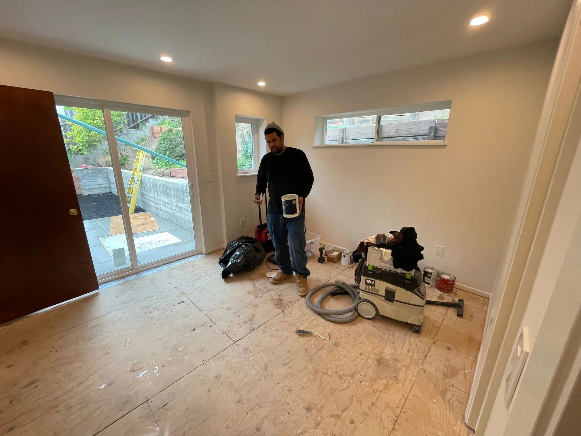 Person standing in a nearly empty room with tools and materials on the floor, near a large window and open door.