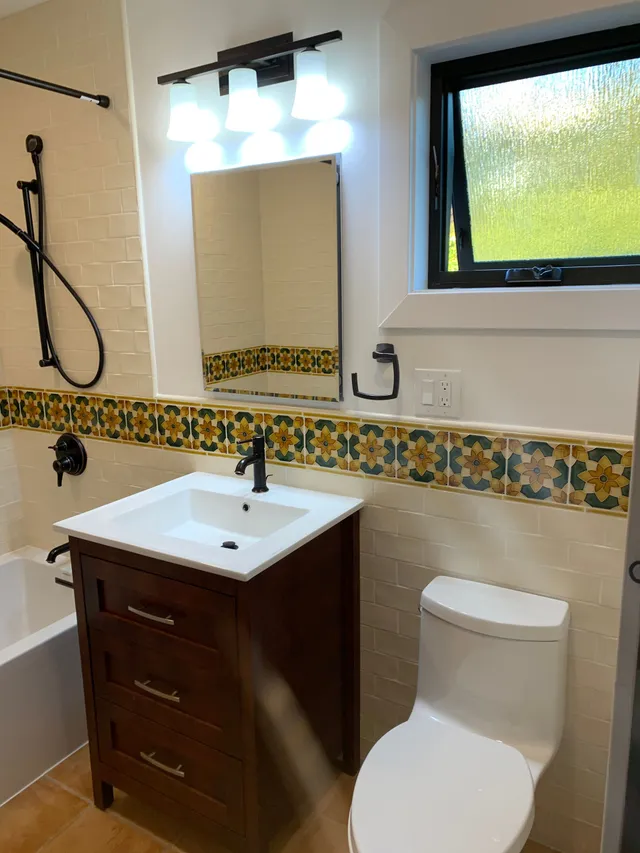 Small bathroom with white sink, wood vanity, toilet, mirror lights, and decorative tile trim.