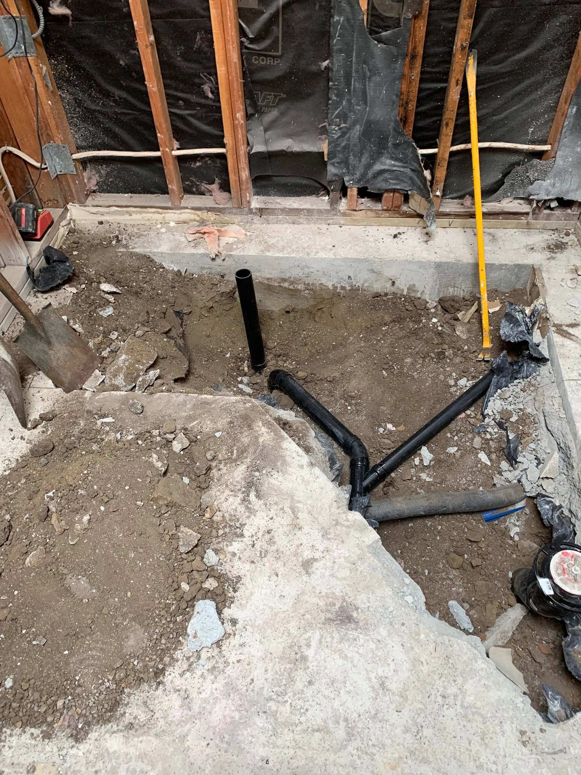 Construction hole in concrete floor with exposed pipes and debris inside a wall-framed room