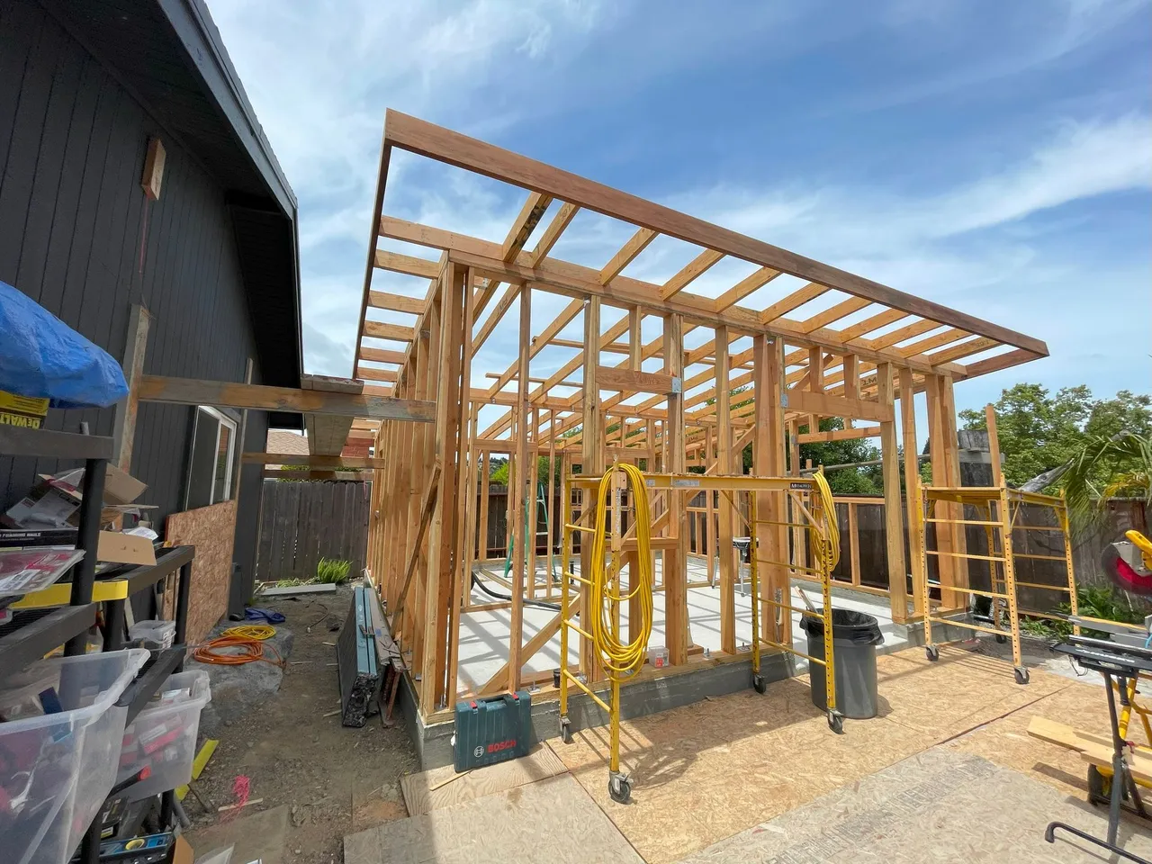 Wood-framed house addition under construction beside a dark building, with scaffolding and lumber on a dirt lot