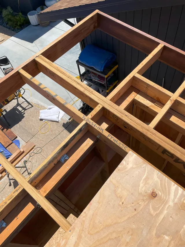 Rooftop under construction with exposed wooden framing, plywood, and a blue tool bag under the roof opening