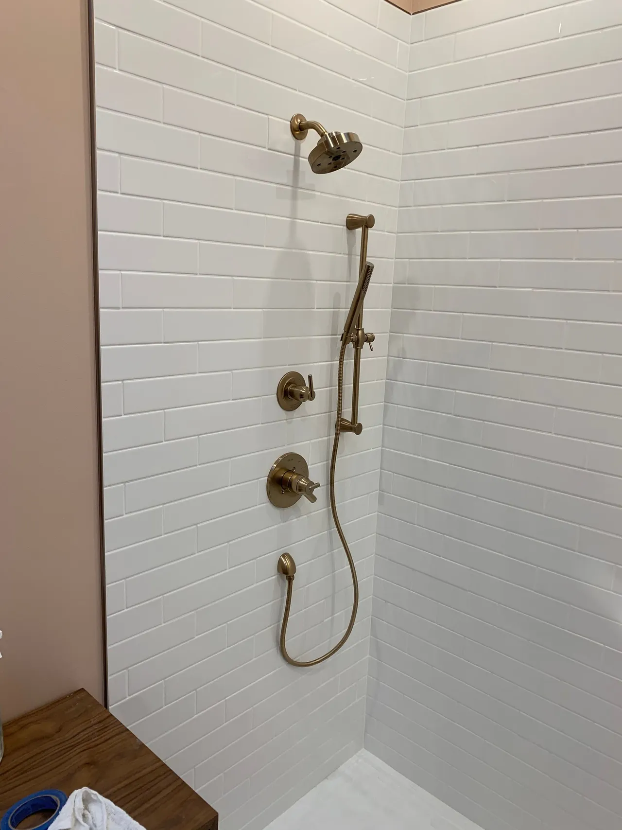White-tiled walk-in shower with brass fixtures, handheld sprayer, and wooden floor outside.