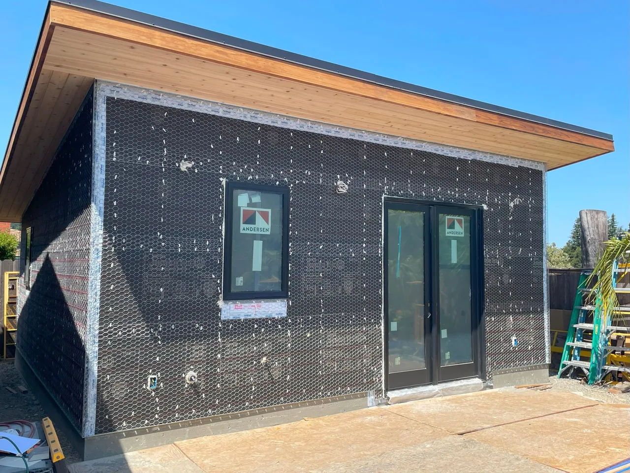 Small modern building exterior with black wrap, wood roof trim, two windows, and a glass door under construction