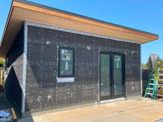 Small modern building exterior with black wrap, wood roof trim, two windows, and a glass door under construction