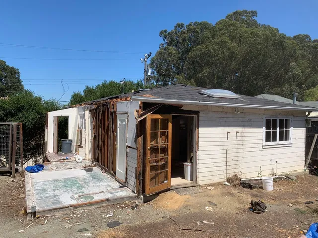 Damaged single-story house with broken doors and a patched roof in a dusty yard under a clear blue sky