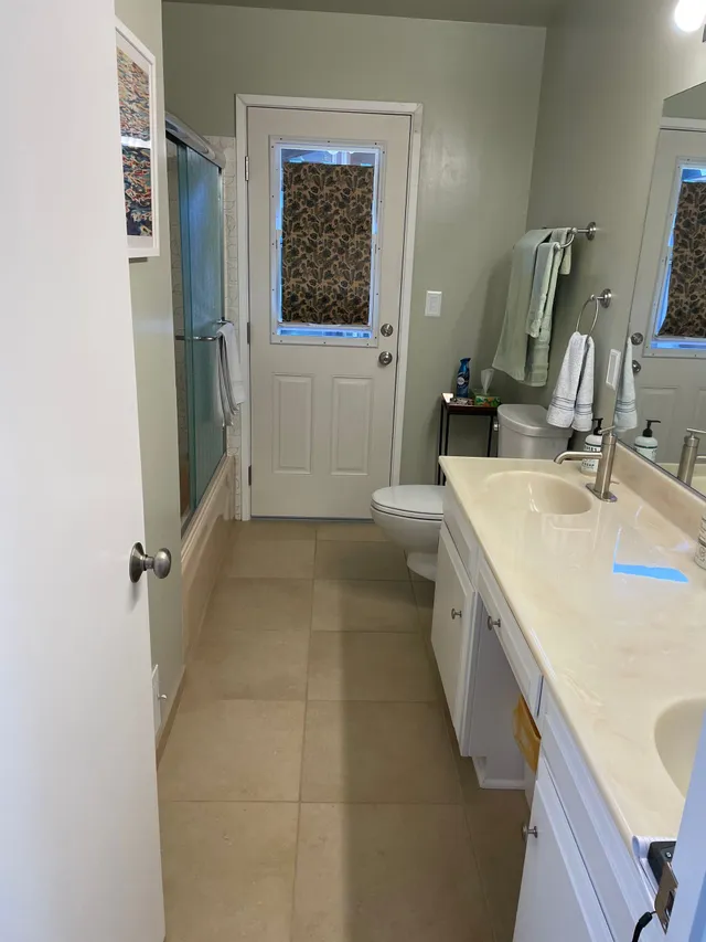 Narrow bathroom with beige tile, white vanity, toilet, and a door with a window at the end.