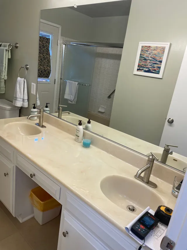 Bathroom vanity with double sinks, large mirror, shower in background, and toiletries on the counter