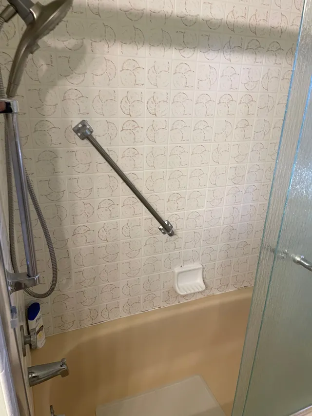 Bathtub shower area with beige tile, glass partition, grab bars, and a handheld shower hose