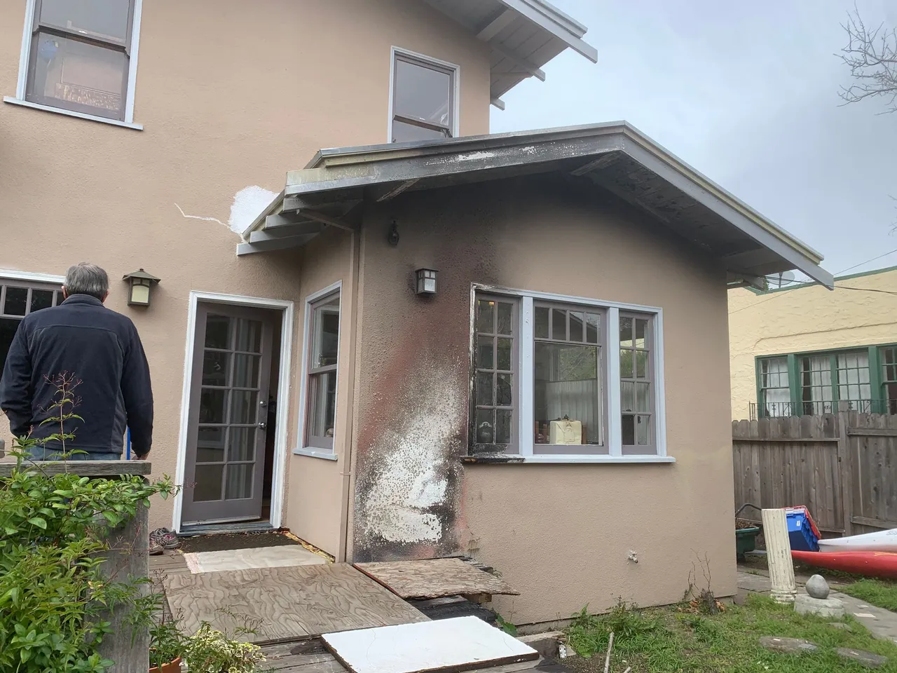 Damaged beige house exterior with broken siding and a person standing by the back door