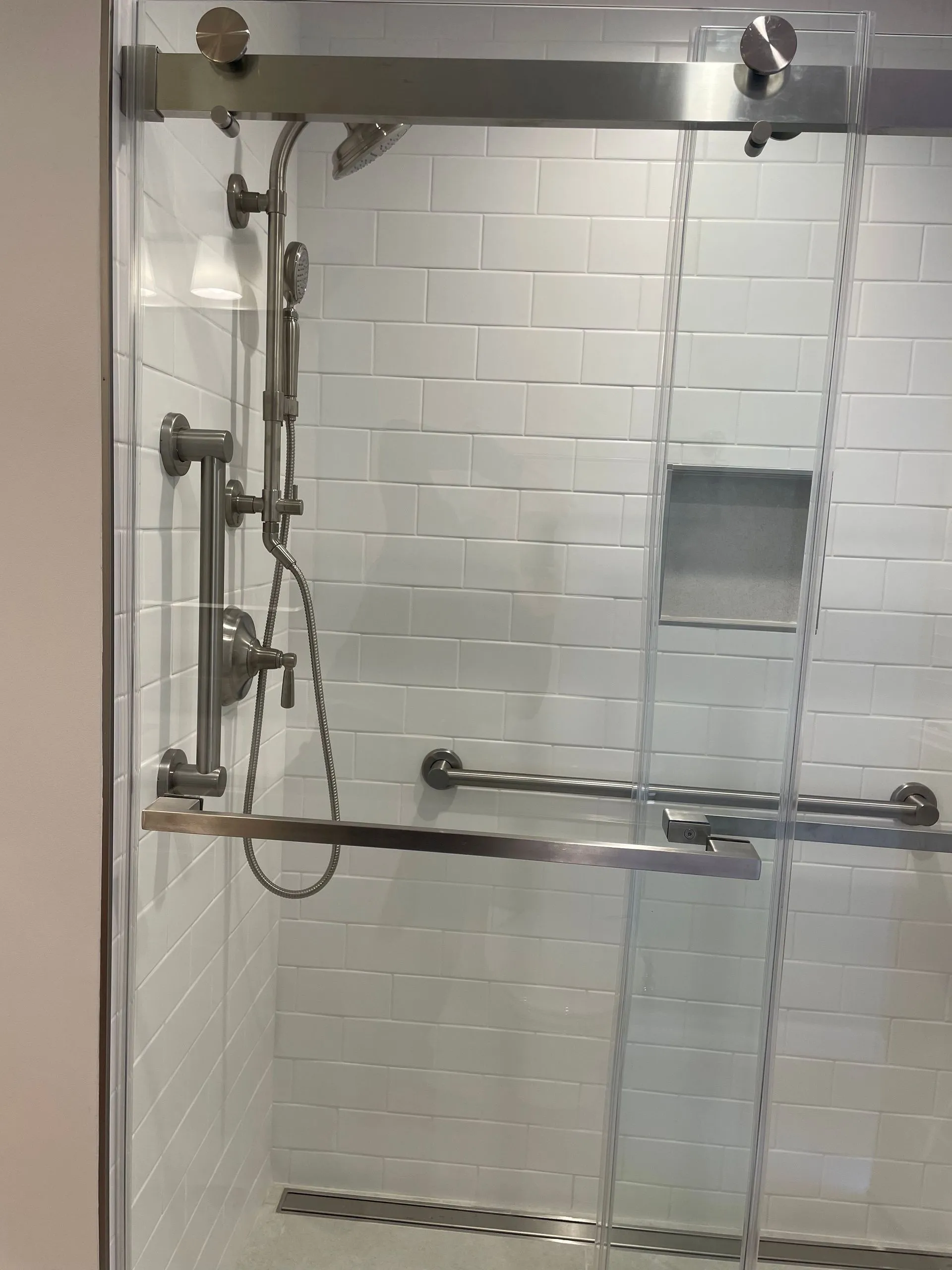 Accessible shower stall with grab bars, handheld shower, and built-in shelf behind glass doors