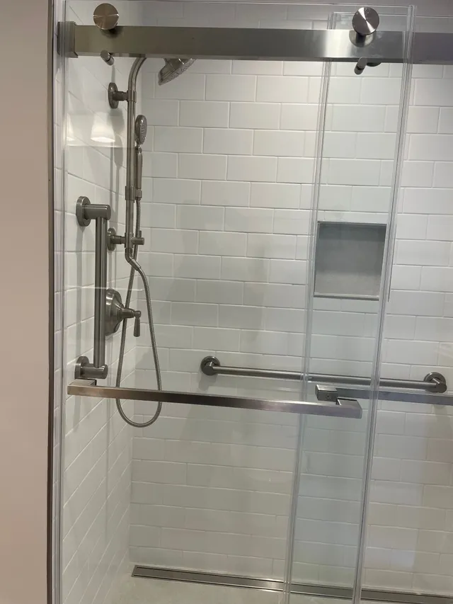 Accessible shower stall with grab bars, handheld shower, and built-in shelf behind glass doors