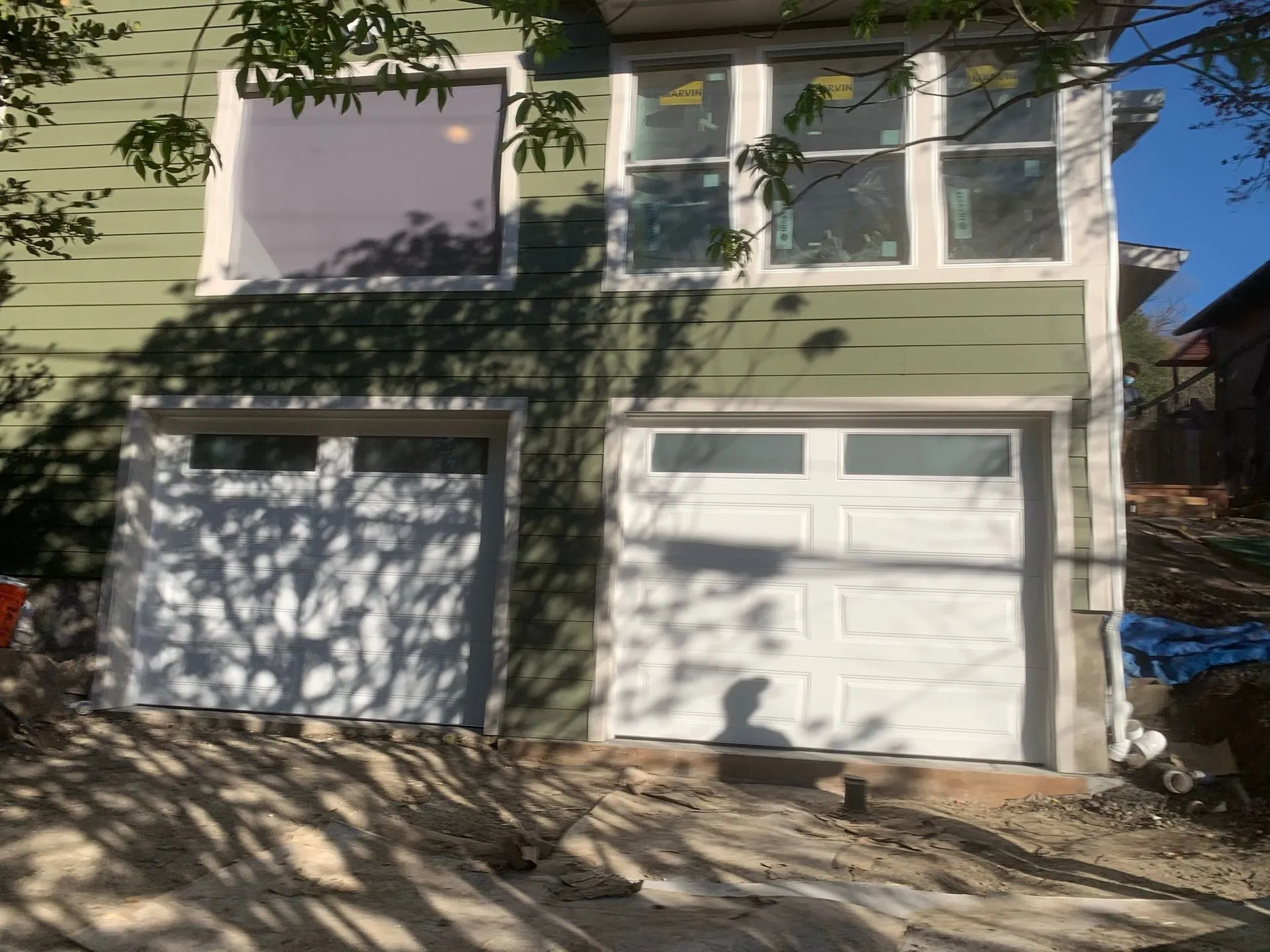 Two-car garage with white doors on a light green house, partly shaded by tree branches
