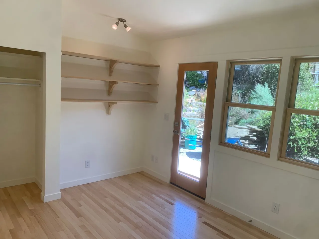 Empty beige room with light wood floor, wall shelves, closet, and windows by a glass door.
