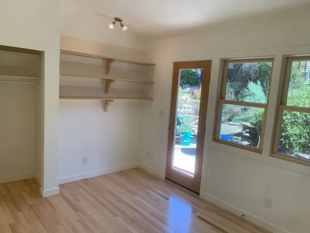 Empty beige room with light wood floor, wall shelves, closet, and windows by a glass door.