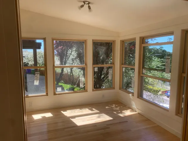 Empty sunlit room with large windows and hardwood floor, showing trees outside.