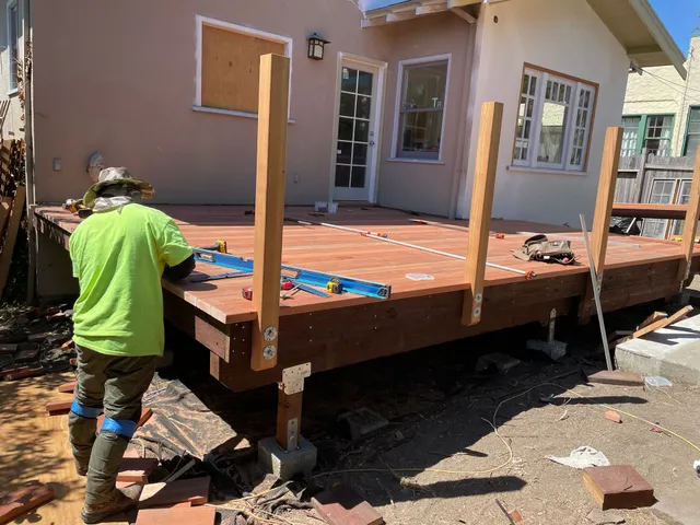 Worker building a wooden deck on a house under construction, with support posts and tools visible