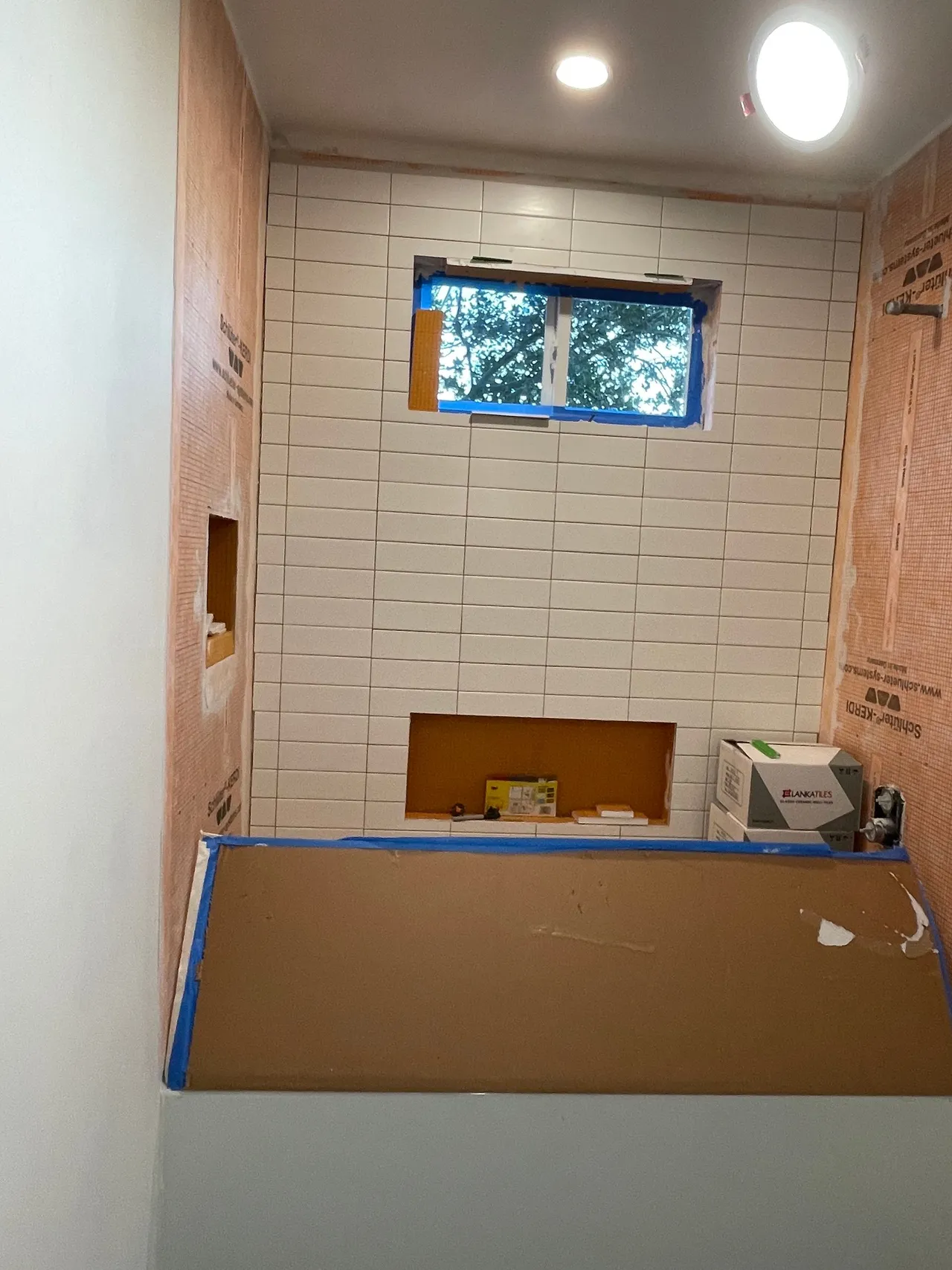 Small bathroom under renovation with tiled walls, a window, and a tub or shower covered by cardboard.
