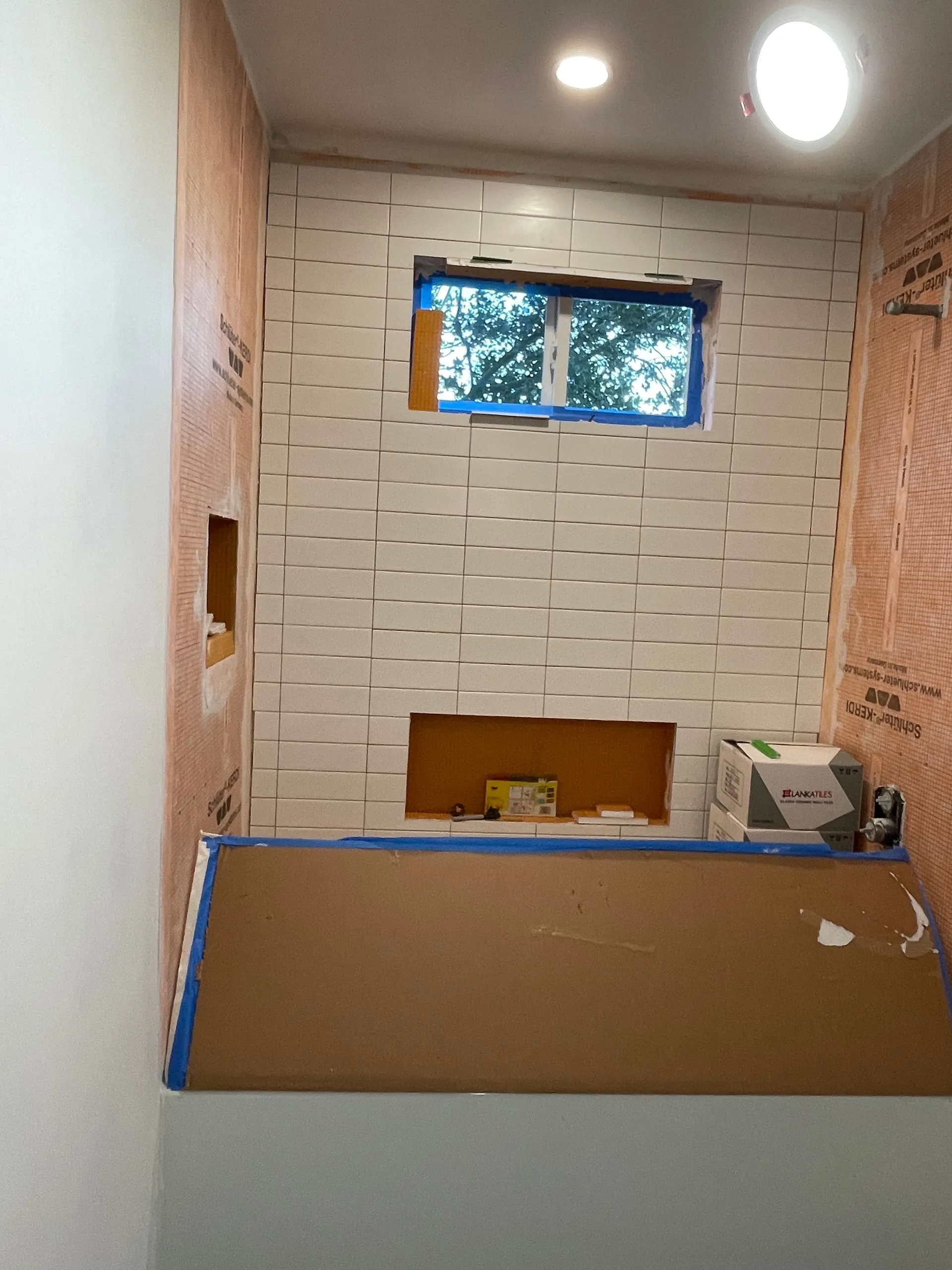 Small bathroom under renovation with tiled walls, a window, and a tub or shower covered by cardboard.