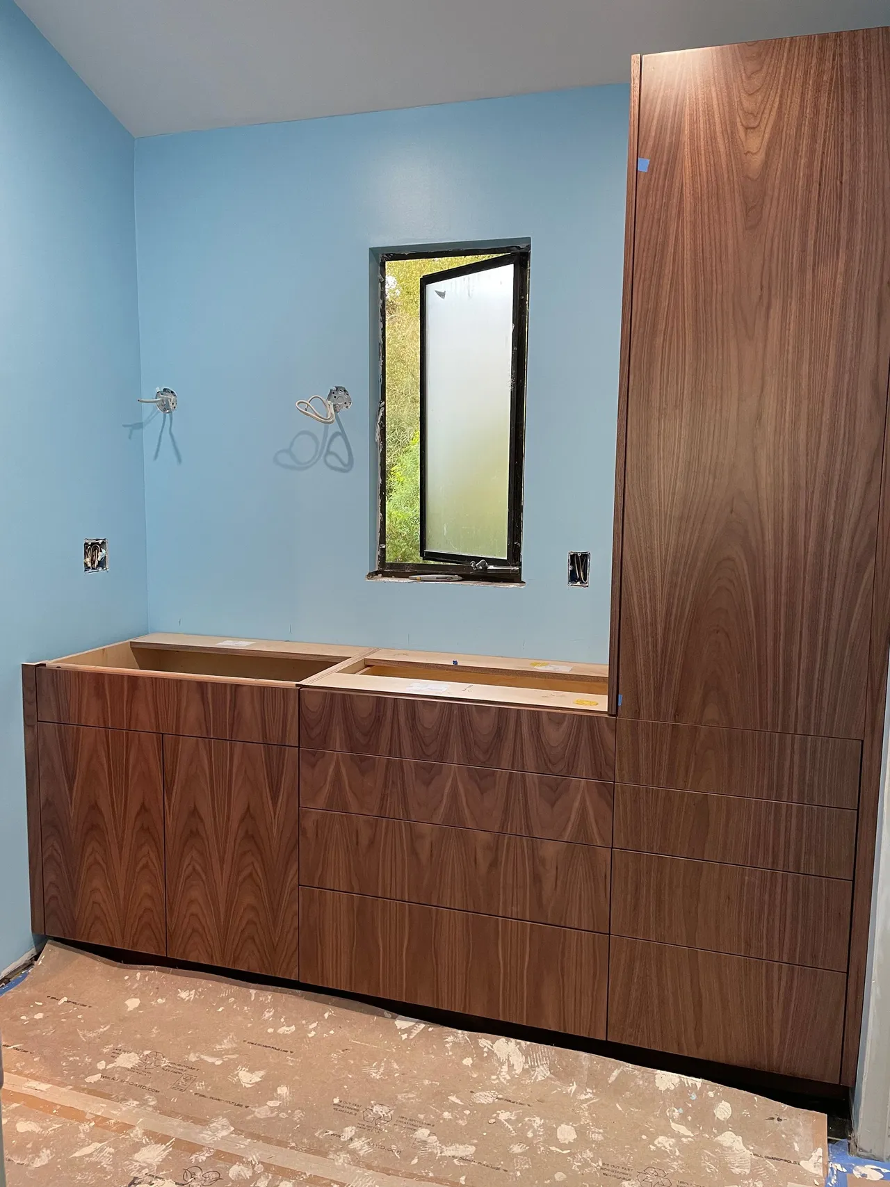 Bathroom vanity under installation with blue walls, wood cabinets, and a narrow vertical window.