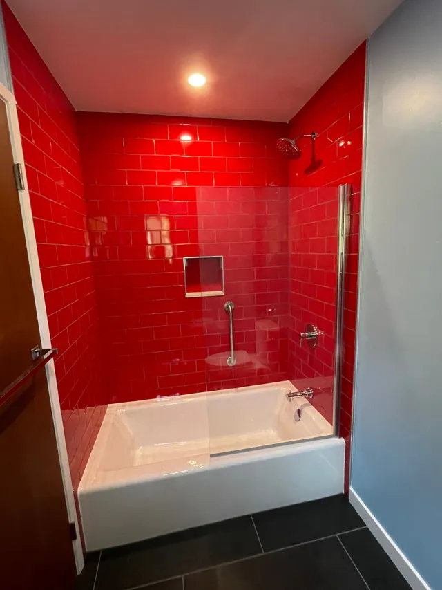 Red-tiled shower alcove with white bathtub, glass panel, and dark floor tiles
