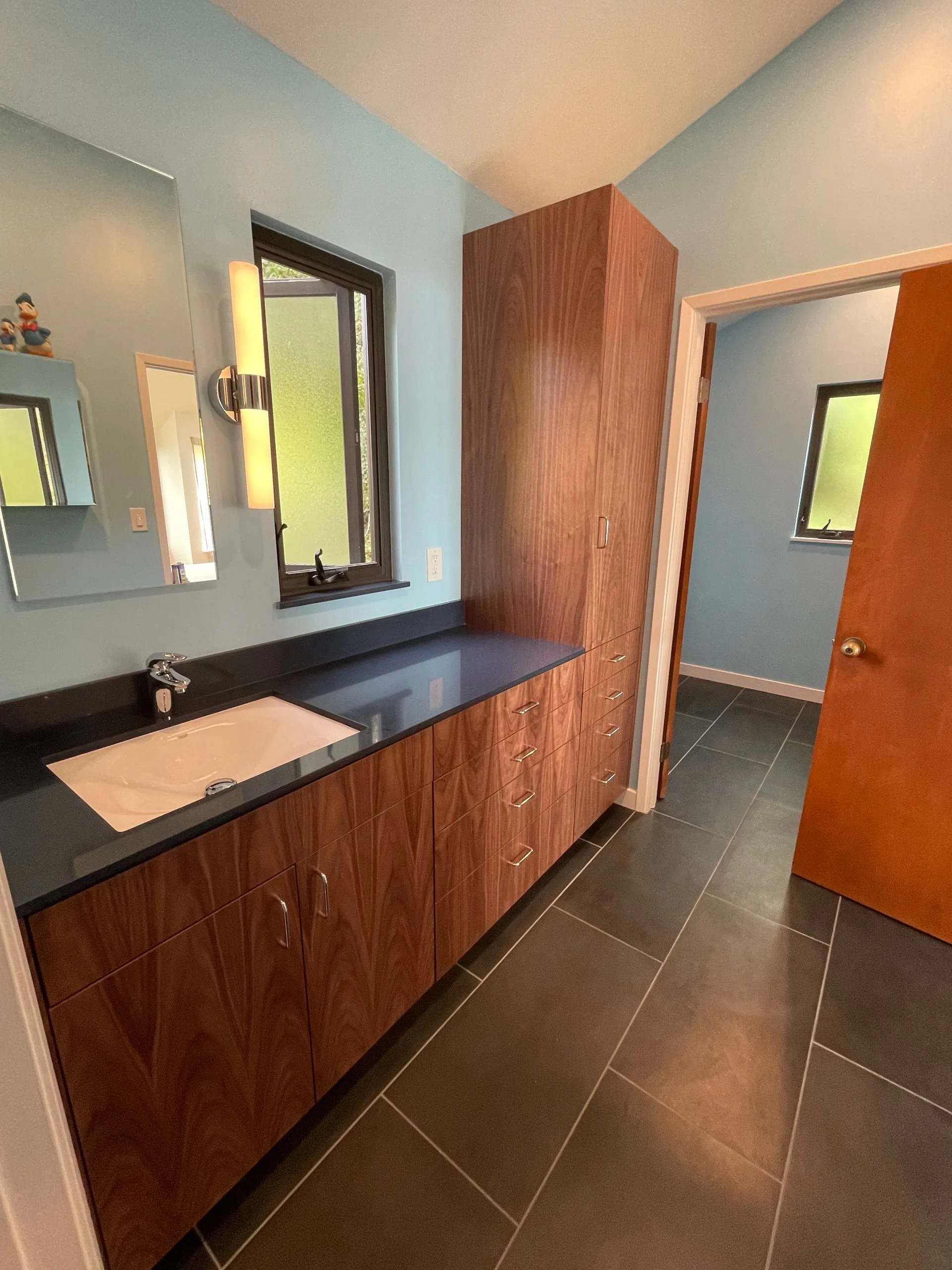 Modern bathroom vanity with wood cabinets, dark countertop, white sink, mirror, and tiled floor