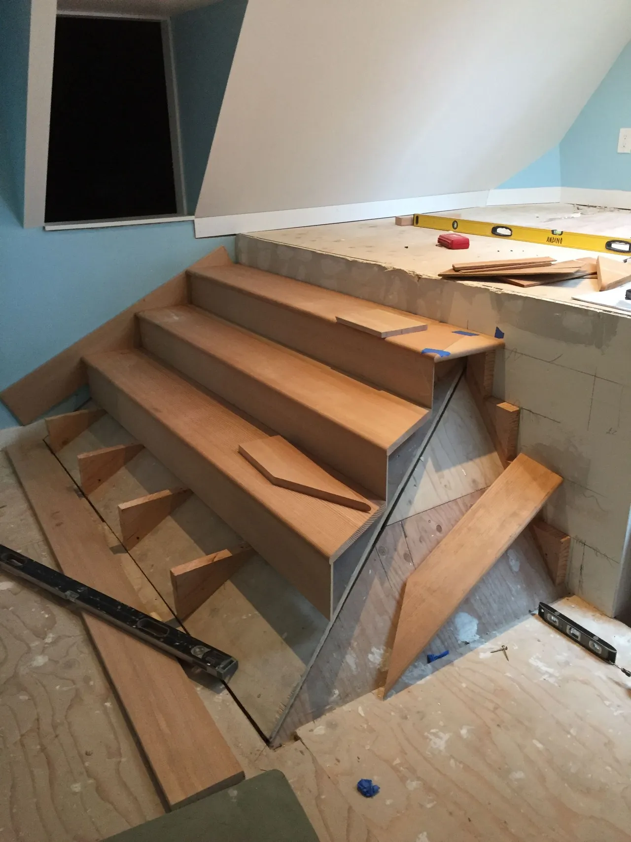 Wooden staircase under construction in a small attic room with unfinished walls and tools nearby