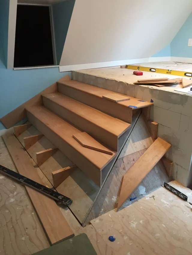 Wooden staircase under construction in a small attic room with unfinished walls and tools nearby