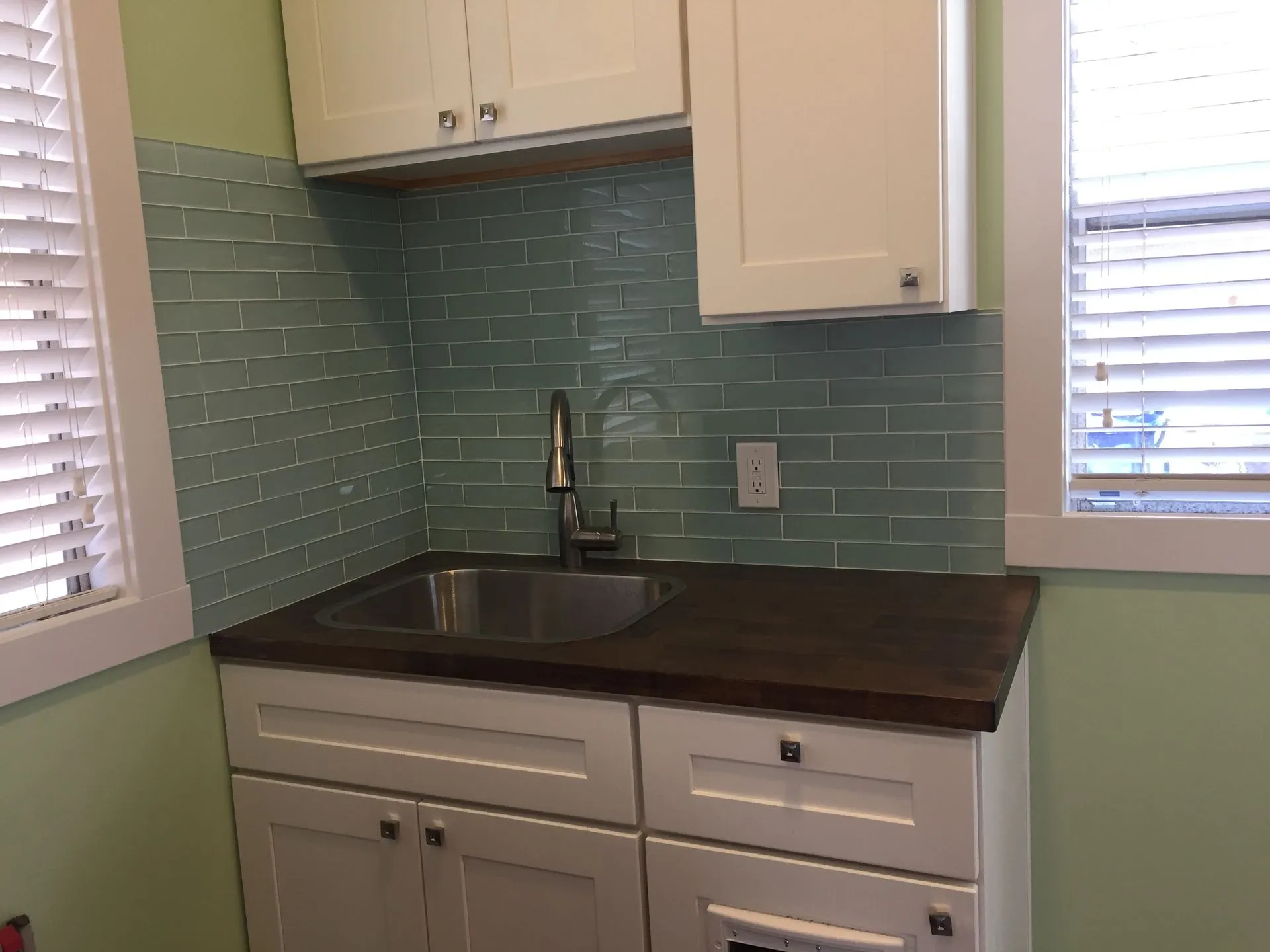 Small kitchen corner with mint tile backsplash, dark countertop, white cabinets, and a sink under cabinets and windows.