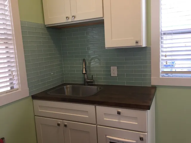 Small kitchen corner with mint tile backsplash, dark countertop, white cabinets, and a sink under cabinets and windows.