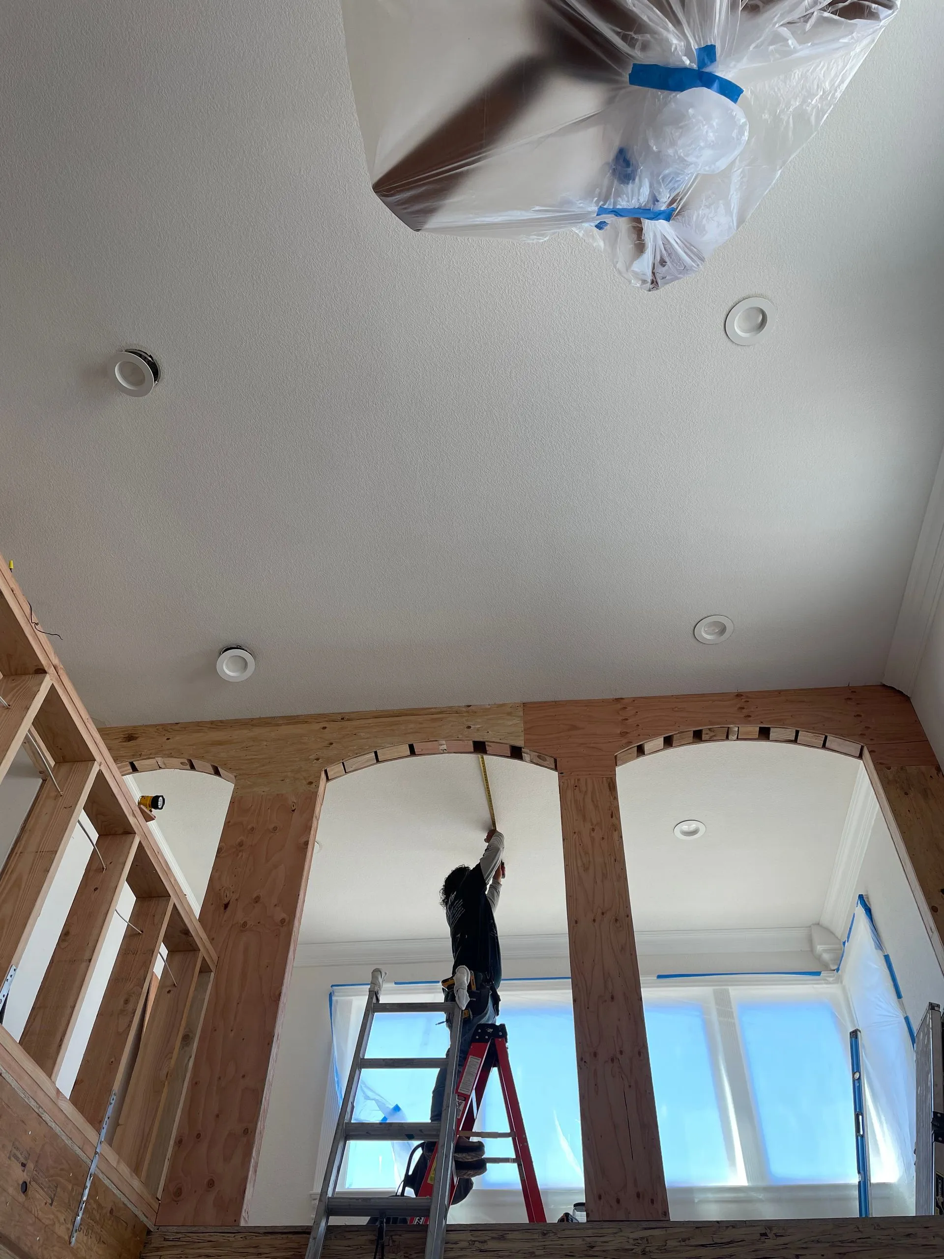 Worker on a ladder installing a ceiling fixture in a room with unfinished walls and windows.