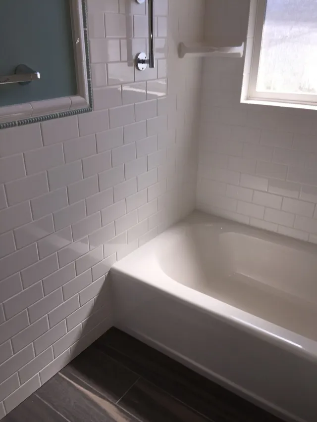 White-tiled bathroom corner with a bathtub, window, and dark wood-look floor.