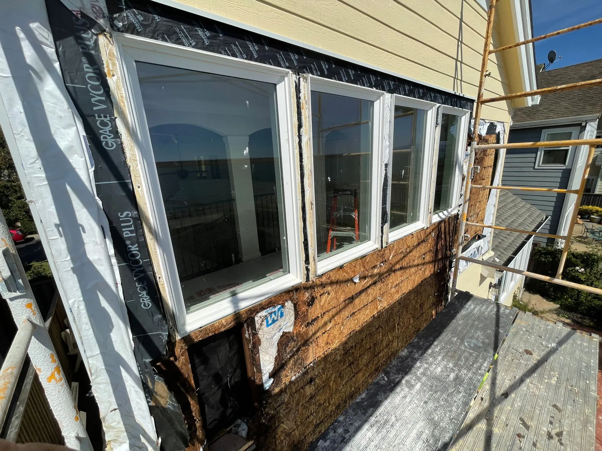 Exterior of a house with white-trim windows and siding under renovation, with scaffolding beside it