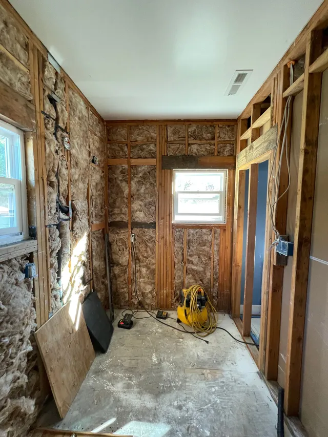Narrow unfinished room with exposed studs, insulation, windows, and a yellow tool on the floor