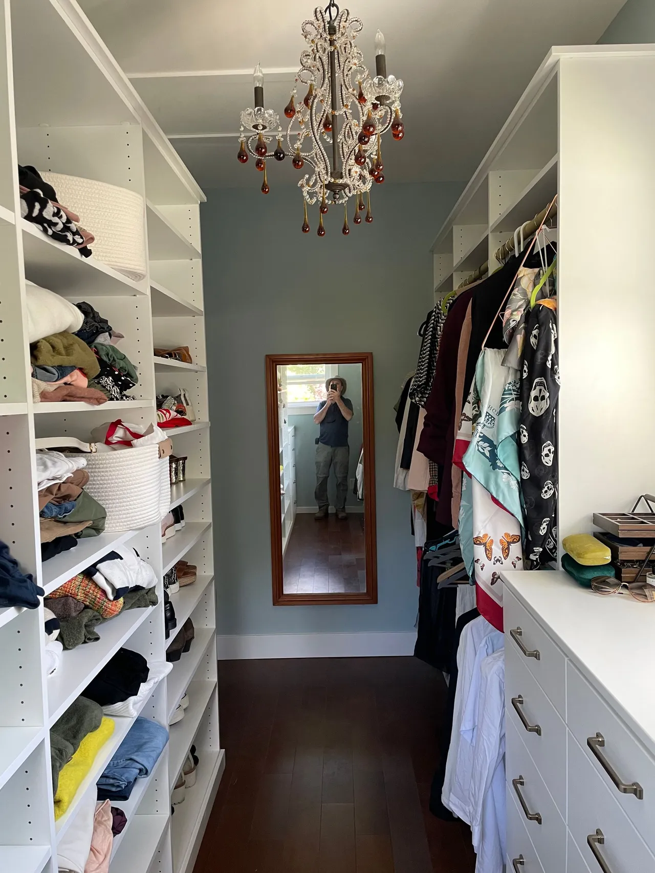 Walk-in closet with white shelves, hanging clothes, and a mirrored door reflecting the room.
